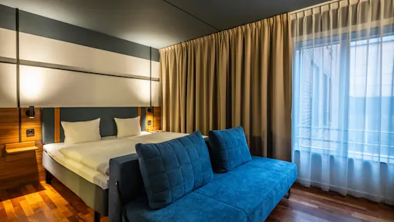  A family room featuring a large double bed and a blue sofa bed. The room has wooden floors, light curtains covering the window, and a modern headboard with built-in bedside tables and reading lights.