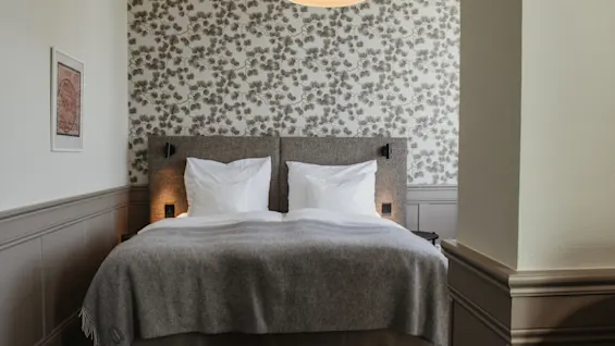 Ett bekvämt hotellrum med en säng med vita kuddar, en grå filt och dekorativ tapet, upplyst av varma sänglampor.