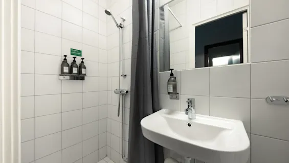 Et lyst badeværelse med bruser og håndvask. Et skilt på væggen lyder SAVE THE PLANET WASH YOUR TOWELS ONLY WHEN NEEDED.