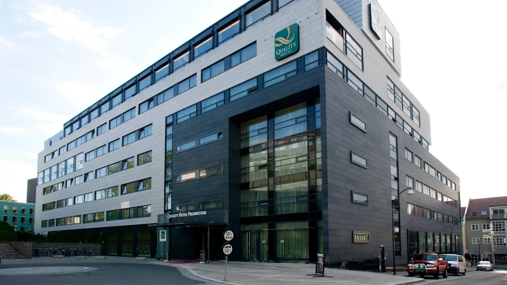A modern hotel building stands with large windows and a mix of gray and white facade. Text: "Quality Hotel Fredrikstad." Situated on a quiet street with parked cars.
