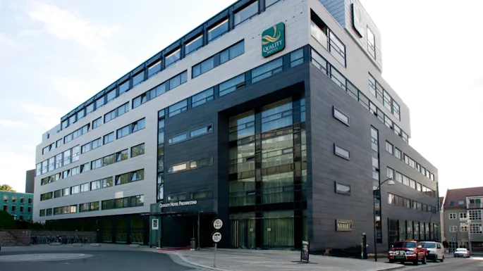 A modern hotel building stands with large windows and a mix of gray and white facade. Text: "Quality Hotel Fredrikstad." Situated on a quiet street with parked cars.