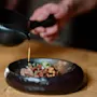 En mørk keramisk skål rummer en garneret ret, mens en hånd hælder sauce fra en sort kande. Miljøet er et træbord.