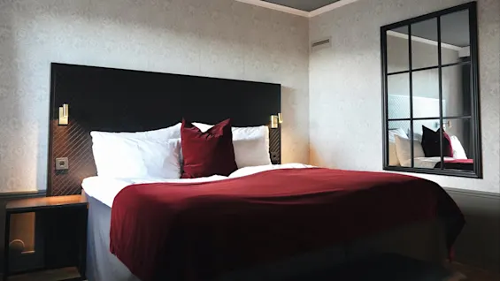 En pent redt seng med rødt pledd og puter står i et stilfullt dekorert rom med et speil på veggen som reflekterer det hele.