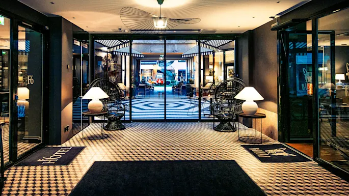 Lobby med mønstret gulv, opplyst av taklamper. To kurvstoler med lamper ved siden av glassdører, leder til et opplyst område. Inngangsmatter viser "F6" og en hundelogo.