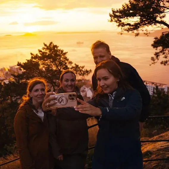 En gruppe venner tar en selfie med en vakker solnedgang over en kystby og vann i bakgrunnen.