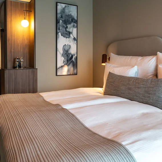 En prydligt bäddad säng med vita lakan och en beige kastfilt står i ett hotellrum. Ett träskåp och abstrakt konst pryder väggen i bakgrunden, med mjuk belysning.