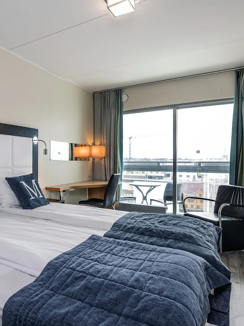 Hotellihuone, jossa on kaksi erillistä vuodetta ja kussakin tyyny "M"-kirjaimella, tarjoaa näkymän kaupunkimaisemaan suurista ikkunoista. Kirjoituspöytä, tuoli ja nojatuoli parantavat toimivuutta.