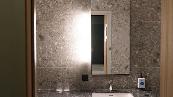 A granite bathroom countertop with a sink below a rectangular mirror reflecting a partially open door. Two glasses and a soap dispenser are on the countertop. Gray tiled walls surround the area.