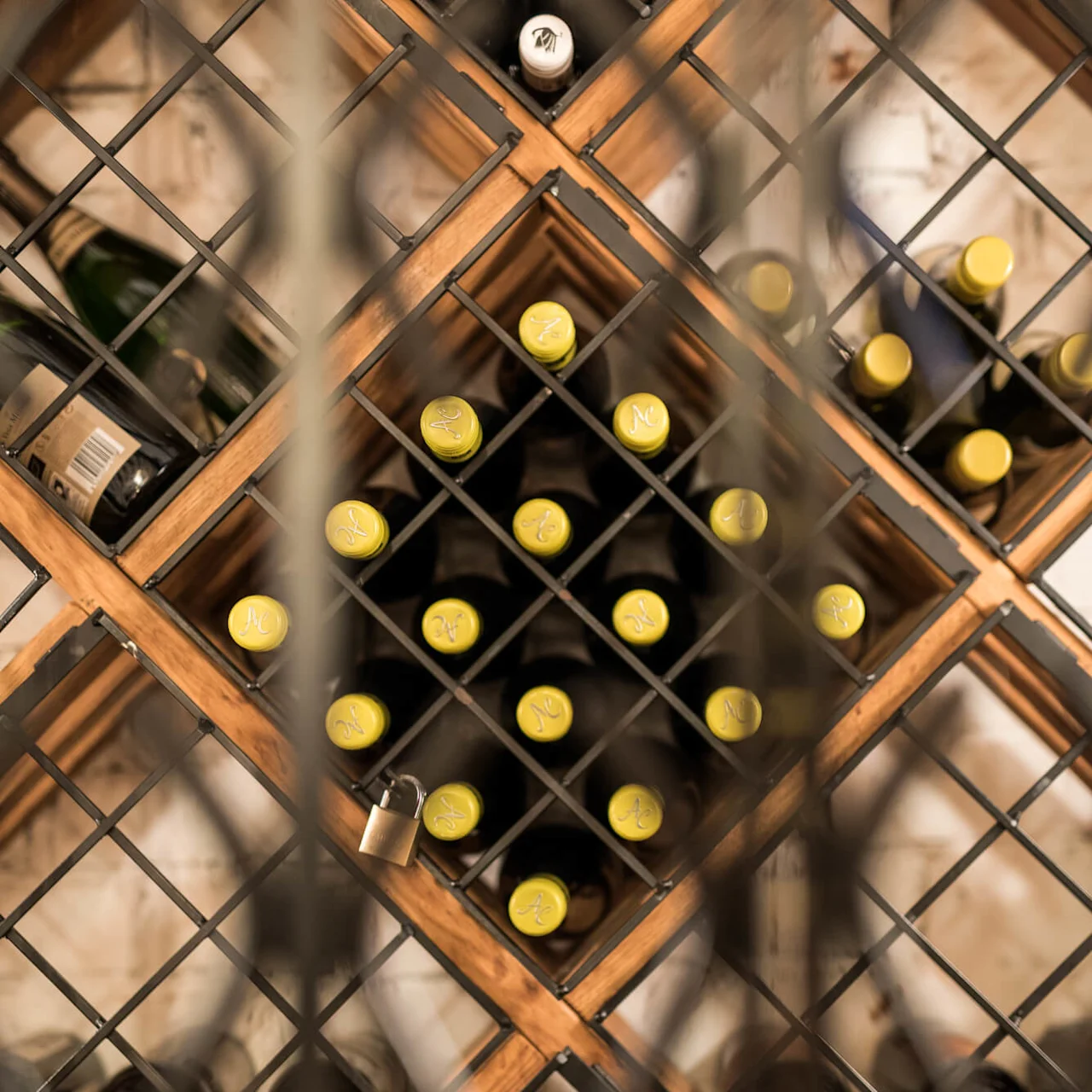 Wine bottles in a wooden rack viewed through a metal lattice, with focus on yellow-capped bottles.