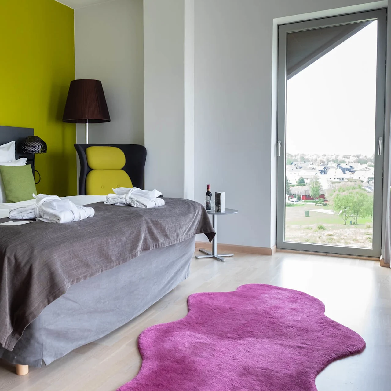 Ett modernt hotellrum har en dubbelsäng med vita lakan, en grön accentvägg och ett fönster med utsikt över byggnader. En rosa matta och en fåtölj kompletterar inredningen.