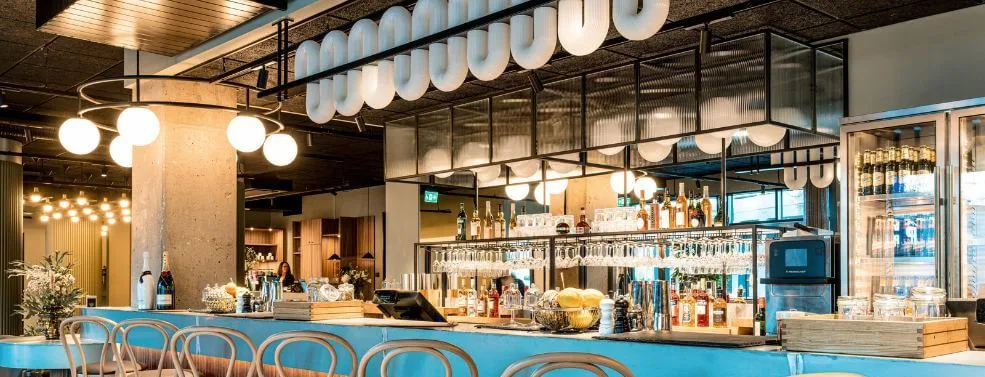 A bar with stools in a modern space, featuring hanging lights and a well-stocked counter with a bartender present.