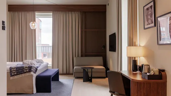 A modern hotel room with a neatly made bed, a sitting area, and a window providing natural light.