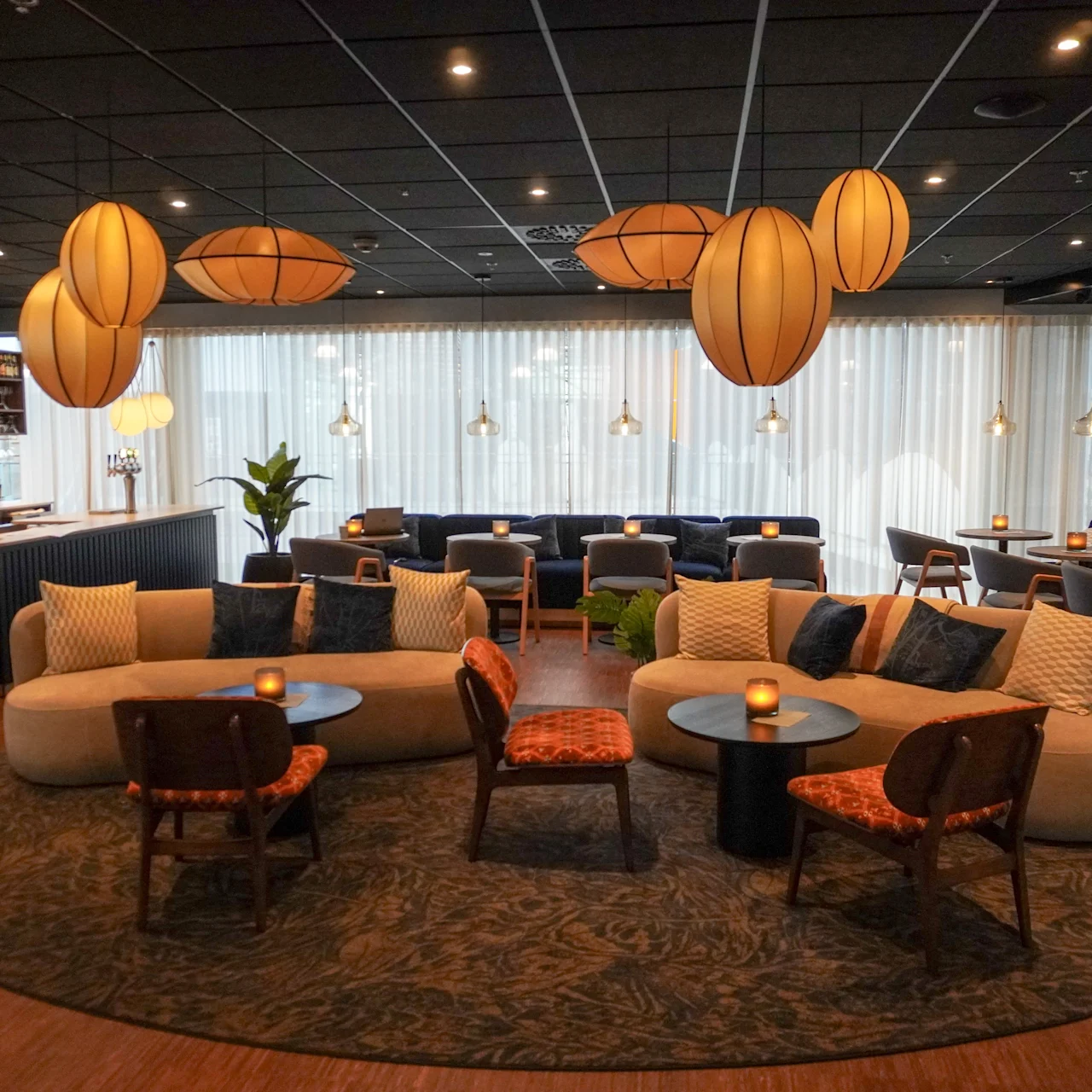 En sittgrupp med moderna soffor och stolar är arrangerad runt små bord. Omgivande hängande lampor ger en mysig atmosfär i en stilfullt inredd lounge.