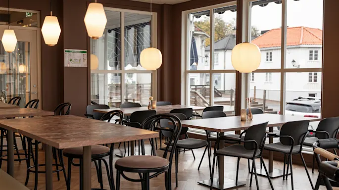 A cozy restaurant interior with various tables and chairs, illuminated by warm pendant lights, offering a view of a street outside.