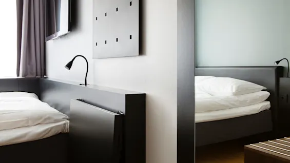To pænt opredte senge med hvidt sengelinned adskilles af en væg i et minimalistisk rum. Vægmonteret TV, læselamper og moderne dekoration giver en elegant, ren atmosfære.