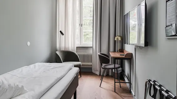 A single bed with white bedding in a tidy room with a desk, chair, and wall-mounted TV near a window with curtains.
