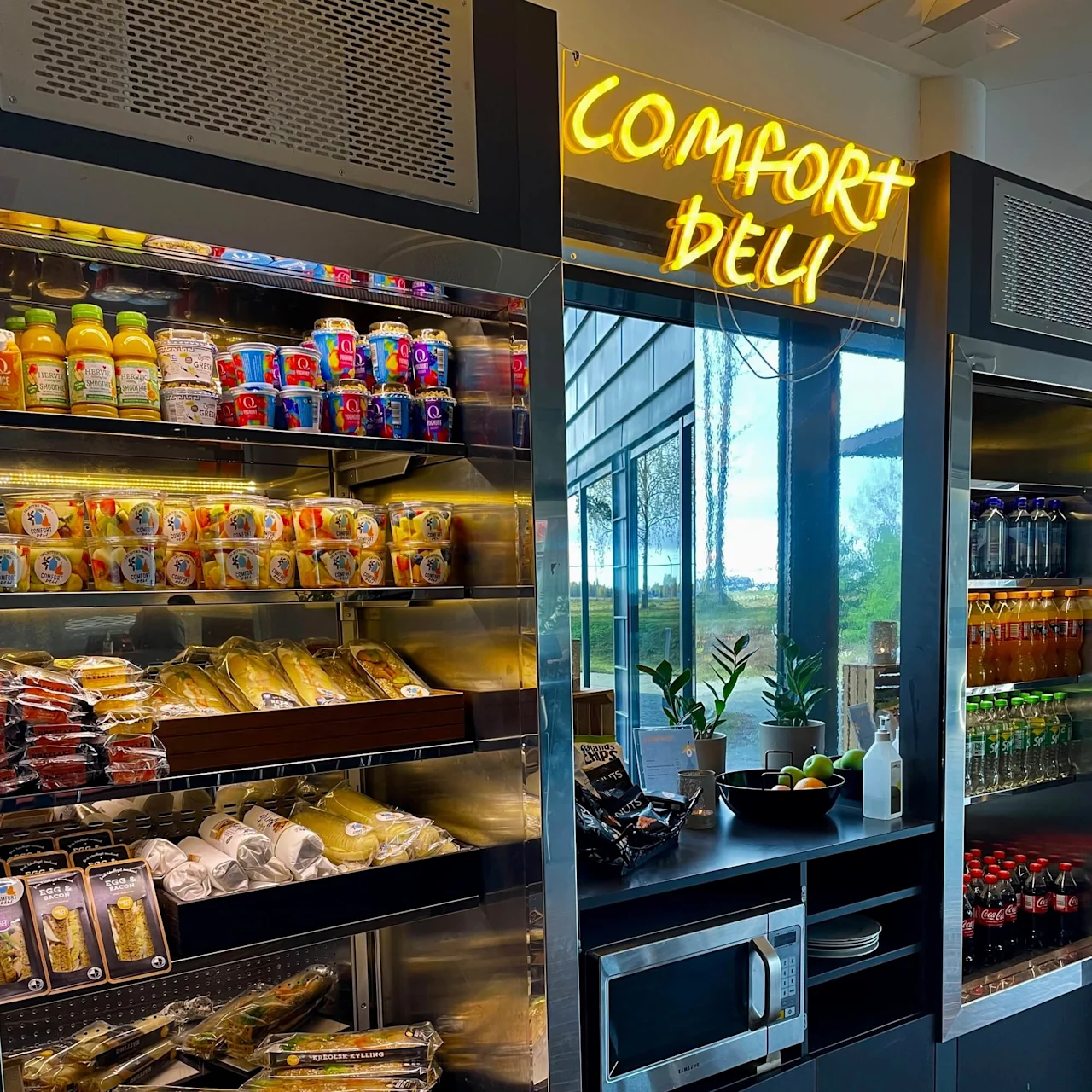 Shelves display sandwiches, juices, and snacks; a neon sign reads "COMFORT DELI." A window shows a garden view; a microwave and fruit bowl sit on the counter.