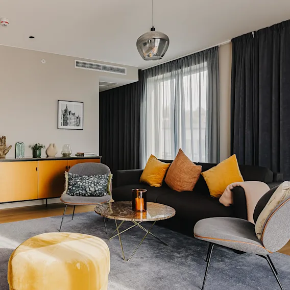 A living room features a black sofa with colorful cushions, a yellow pouf, a small round table, and a media cabinet, surrounded by dark curtains and wooden flooring.