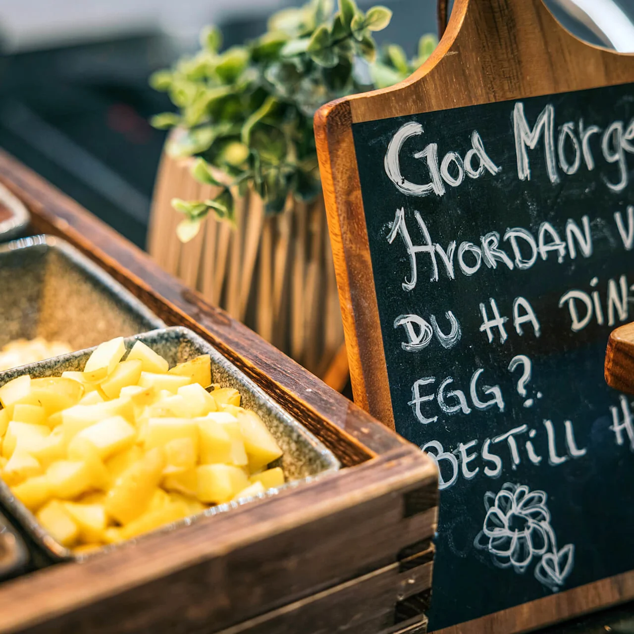 Skålar med hackad potatis och andra ingredienser står bredvid en griffeltavla där det står "God Morgen. Hvordan vil du ha dine egg? Bestill her," med en växt i bakgrunden.