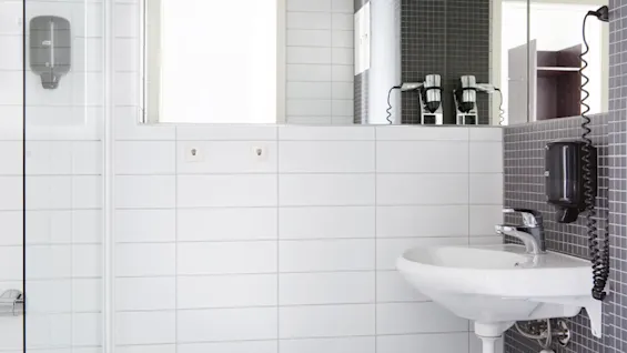 En hvit vask er montert på grå fliser, under et speil som reflekterer en hårføner. Såpedispensere er festet til venstre. Sterkt vinduslys lyser opp det flislagte badet.