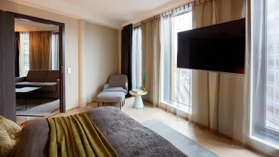 A bedroom features a neatly made bed with a green blanket and connects to a sitting area with a sofa. A television is mounted on the wall, near large windows with curtains.