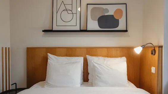 Viihtyisä hotellihuone Home Hotel Uppsalassa siististi pedatulla sängyllä, puisella sängynpäädyllä ja abstraktilla taiteella yläpuolella.