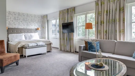 A hotel room features a large bed with a tufted headboard and bedside lamps, adjacent to a sofa and round coffee table. Windows with floral curtains provide natural light.
