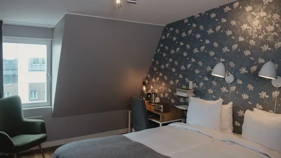 A neatly made bed with white linens beside a patterned floral wall. Adjacent, a desk with a lamp and books sits under angled lighting, and an armchair is near the window.