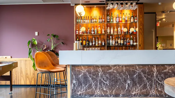 A marble-topped bar with two brown stools stands before a backlit shelf displaying various bottles. A plant sits nearby against a purple wall, creating a modern setting.