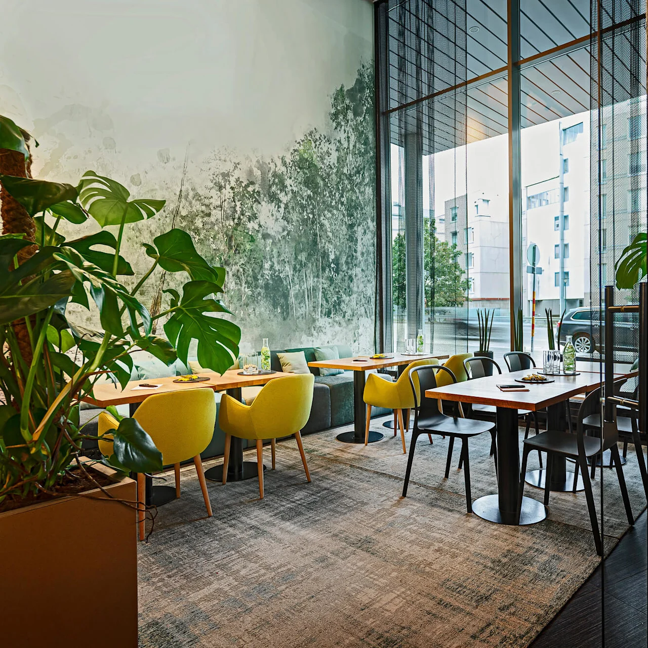 Tables with green and black chairs are situated in a modern restaurant with large plants and a glass wall, overlooking urban buildings.