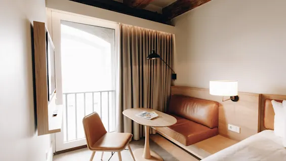 Ett mysigt hotellrum med säng, bord, stol och bänk vid balkongdörr, skapar en ljus och inbjudande atmosfär.