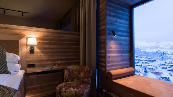 A cozy wooden room features a bed, a patterned armchair, and a large window seat with a snowy mountain view.