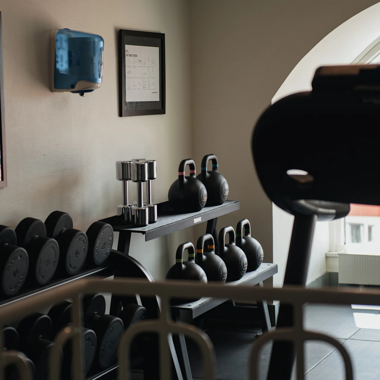 Weights rest on racks; a row of dumbbells and kettlebells are organized neatly. Nearby is a large window providing natural light, enhancing the gym's serene ambiance.