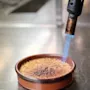 A kitchen torch caramelizes sugar atop round crème brûlée in a brown dish. The dish sits on a metal counter in a kitchen setting.