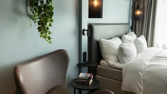 Et koselig hotellrom med seng, hvit sengetøy, komfortabel lenestol og hengende plante, opplyst av stilige vegglamper.