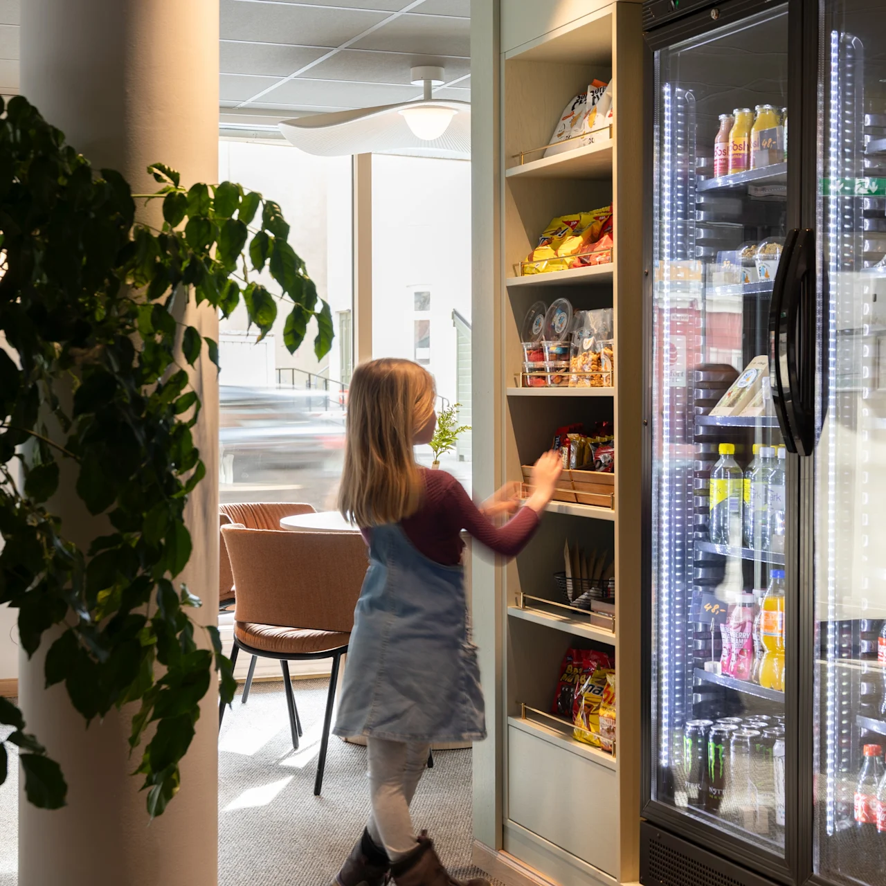 Lapsi kurottaa hyllyiltä ruokatuotteita, viereisessä jääkaapissa on juomia. Tilassa on kirkas valaistus, suuri kasvi ja tuoleja taustalla.