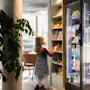 Ett barn sträcker sig efter föremål på snacks-hyllor bredvid ett kylskåp fyllt med drycker. Miljön är ett väl upplyst rum med en stor växt och stolar i bakgrunden.