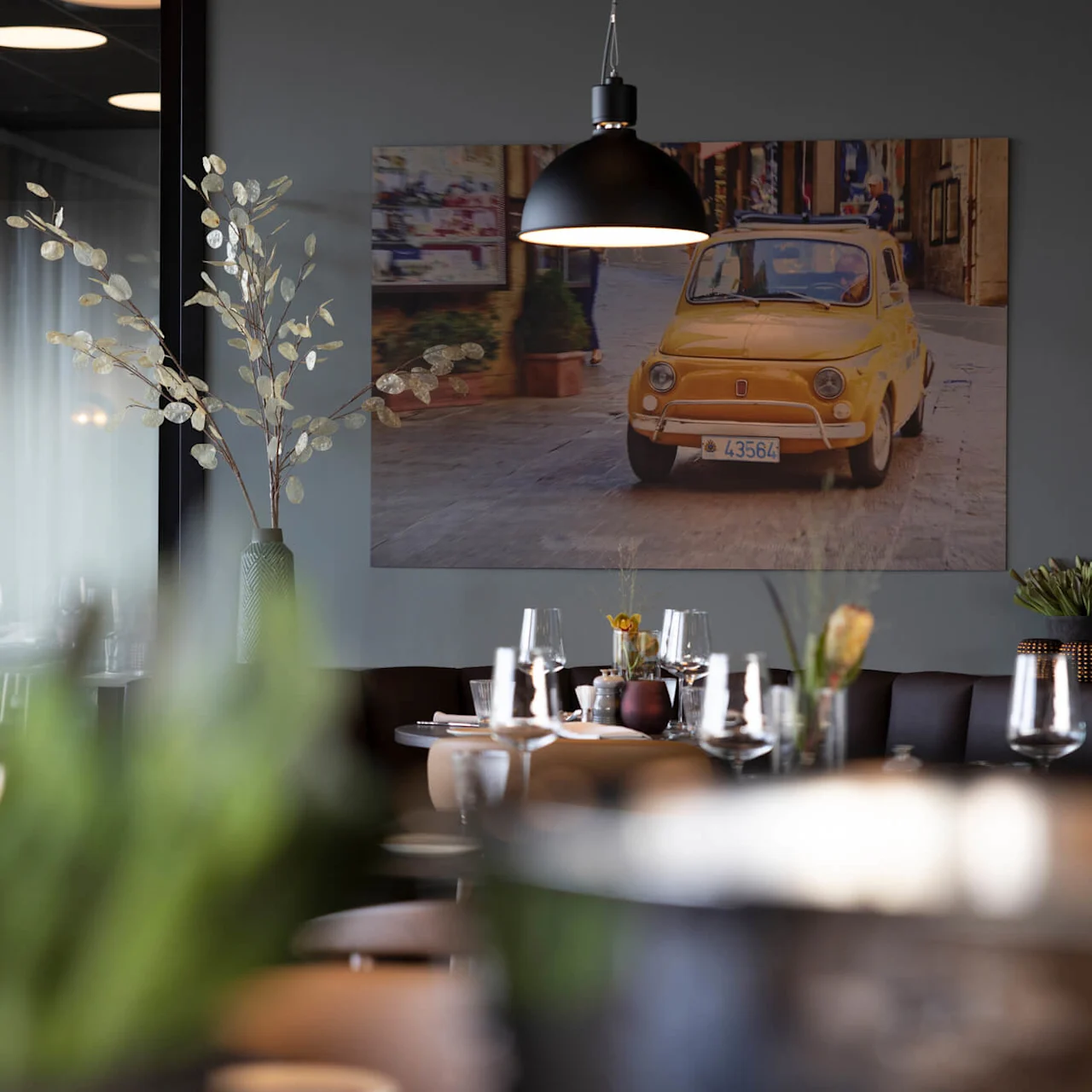 A dining room with a yellow vintage car depicted in a framed photograph, elegant table settings, and modern decor, suggesting an upscale restaurant ambiance.