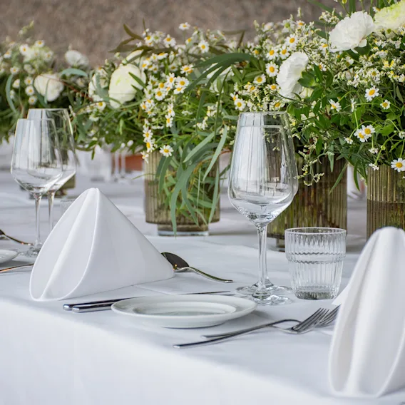 Et vakkert dekket bord med hvite duker, brettede servietter, elegant glass og frodige blomsterarrangementer med hvite blomster.