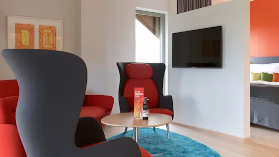 Två moderna stolar och ett litet runt bord med en vattenflaska och en skylt som lyder "GET ME SOMETHING TO EAT" står bredvid en monterad TV i ett stiligt rum.