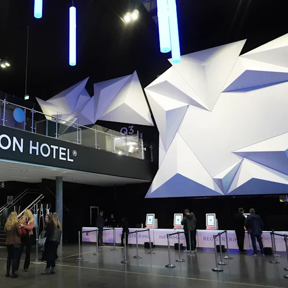 Clarion Hotel sign above a modern reception area with geometric wall design. People are standing by registration kiosks. Sign reads "INFORMASJON REGISTRERING EVENT ARENA."