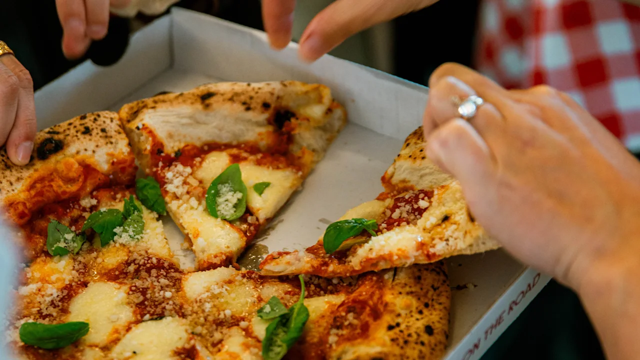 En pizza med smält ost och basilika delas av flera händer från en låda, omgiven av människor i en avslappnad inomhusmiljö.