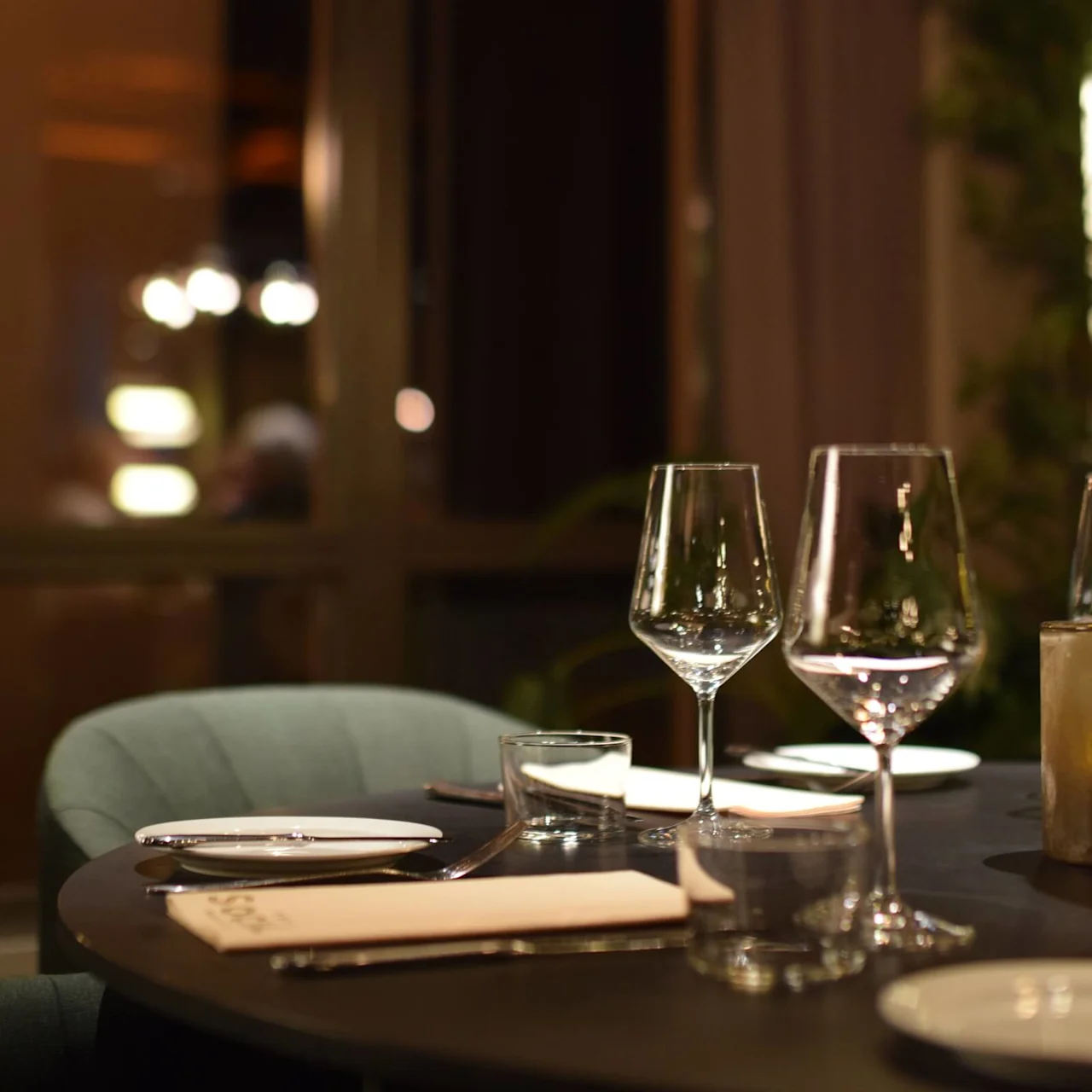 Et bord dækket med tomme vinglas, tallerkener og bestik i en blødt oplyst, elegant restaurant. Et stort vindue og slørede lys giver en varm, hyggelig stemning.
