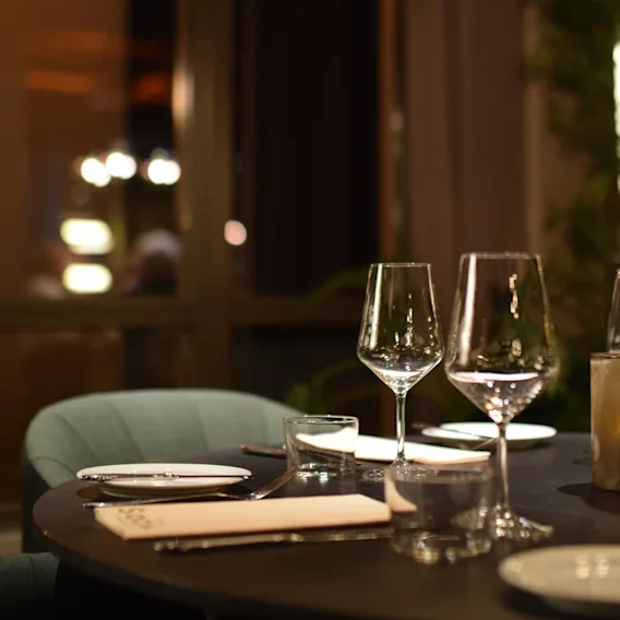Et bord dekket med tomme vinglass, tallerkener og bestikk i en mykt opplyst, elegant restaurant. Et stort vindu og uskarpe lys gir en varm, koselig atmosfære.