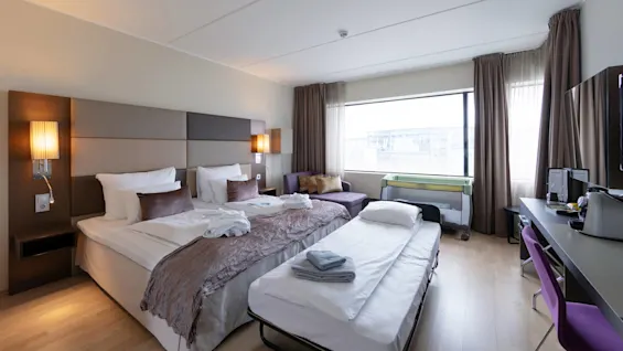 A neatly arranged hotel room features two beds with crisp white linens, a purple chair by a sleek desk, and a comfortable sofa near a large window. Folded towels on beds.