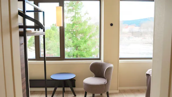 A bright room with a large window overlooking green trees, featuring a cozy chair and a small table.