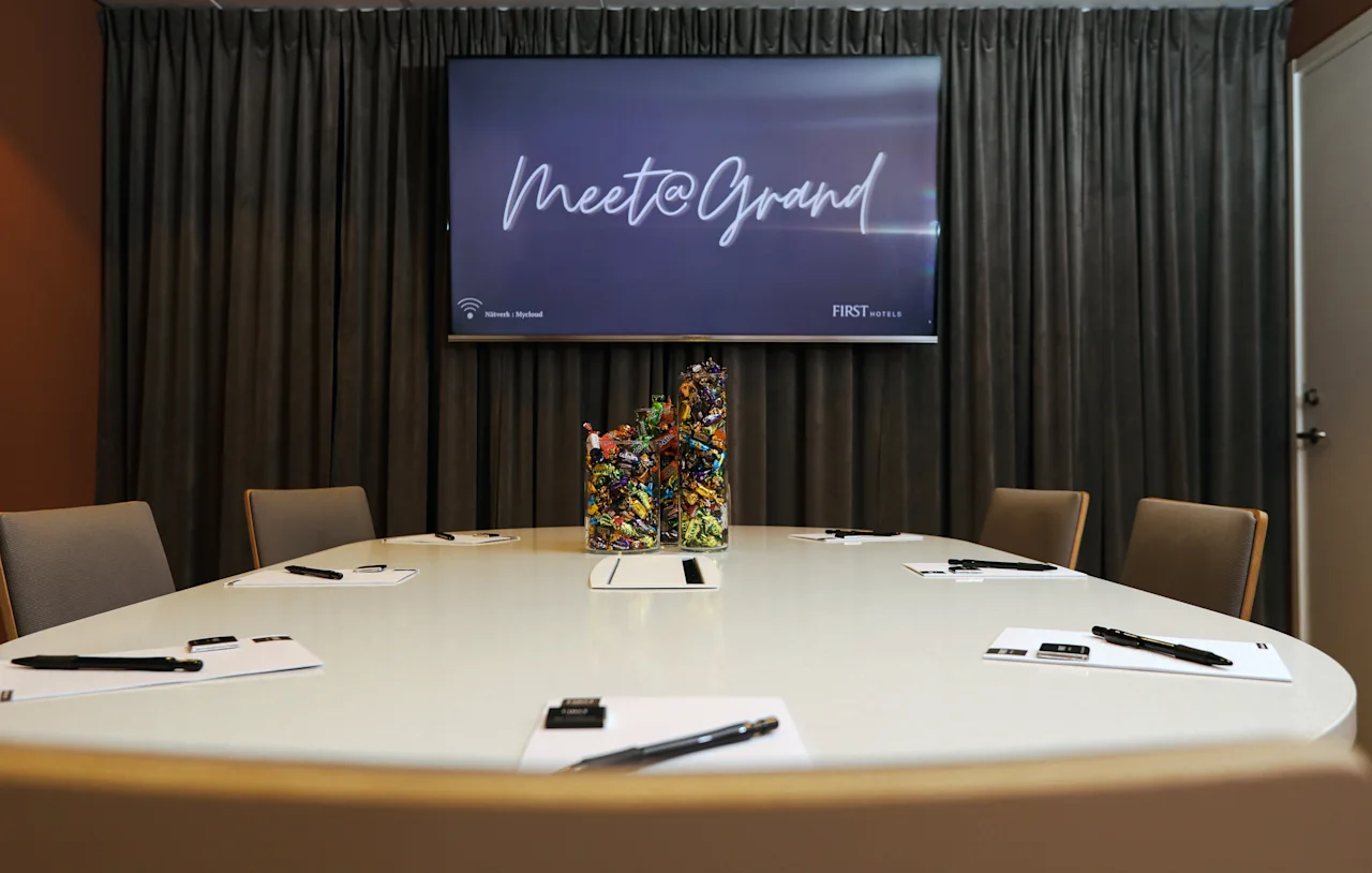 En konferensrum med ett runt bord med anteckningsblock och pennor, centrerat med en färgglad godishållare. En skärm visar "Meet@Grand" mot ett draperat bakgrund.