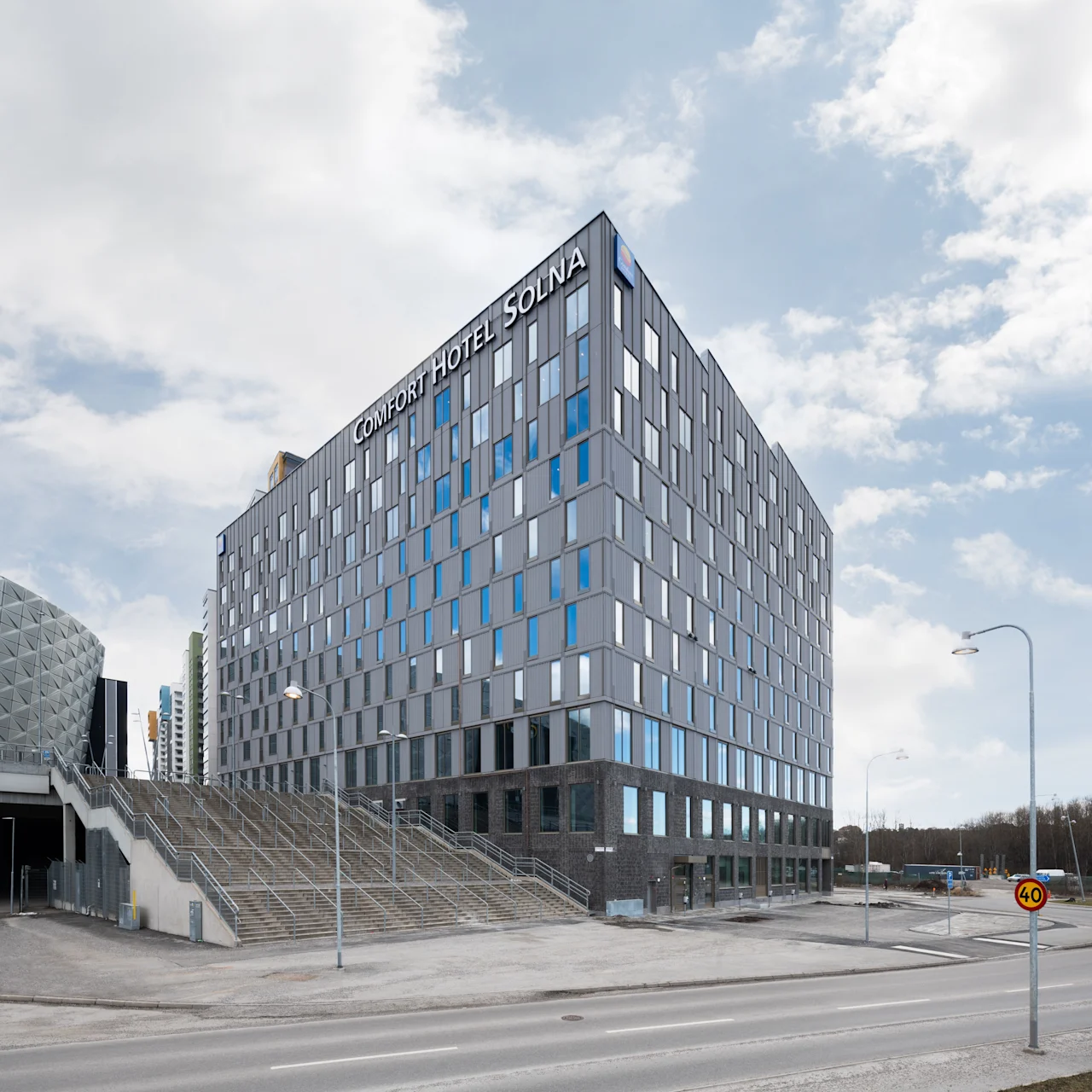 Korkea, moderni rakennus nimeltään "COMFORT HOTEL SOLNA" seisoo näkyvästi kaupunkikadulla, yläpuolella pilvinen taivas ja lähellä kaupunkirakennelmia.