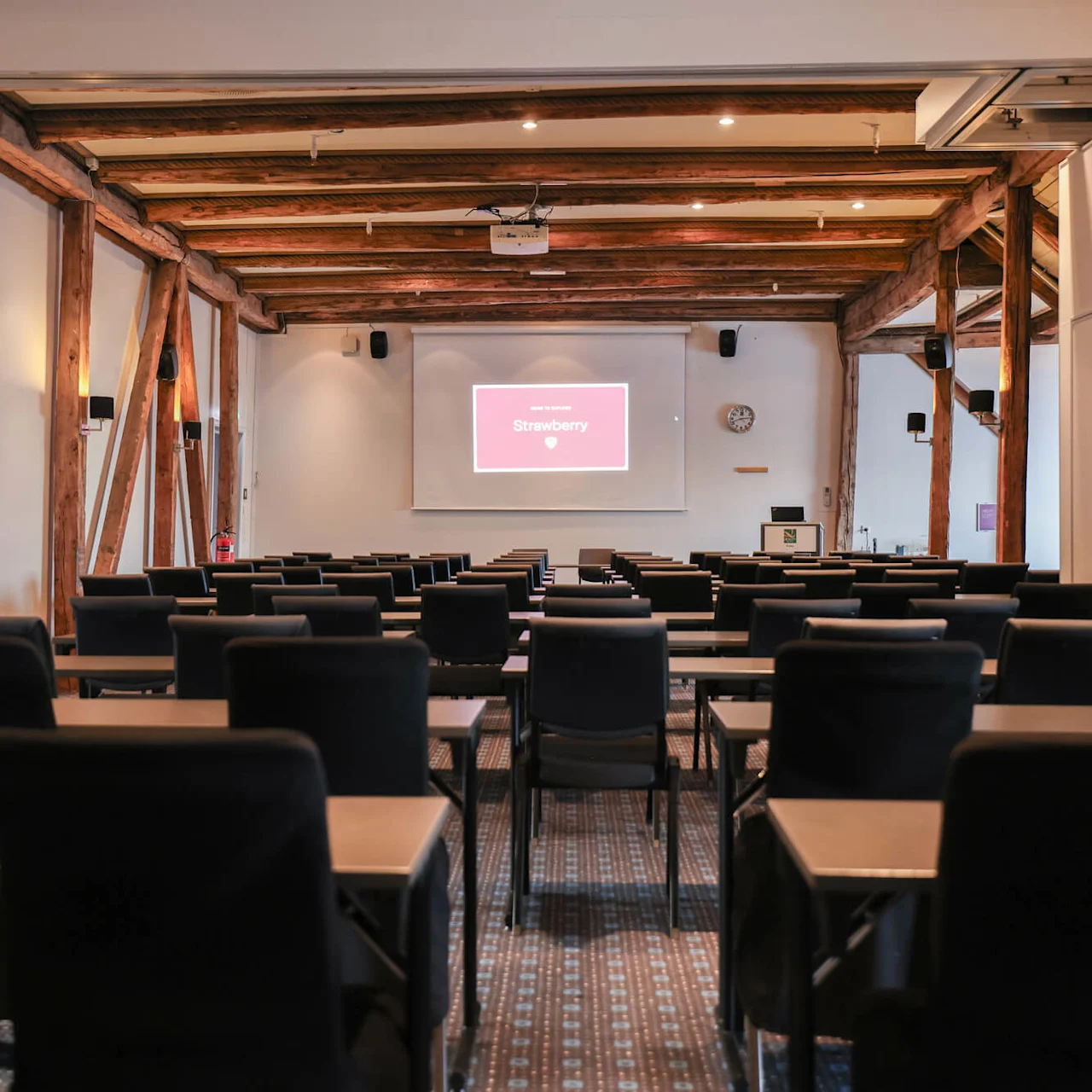 Rows of black chairs face a large screen displaying "Strawberry" in a rustic conference room, with exposed wooden beams and overhead lights. A clock and a podium are visible.