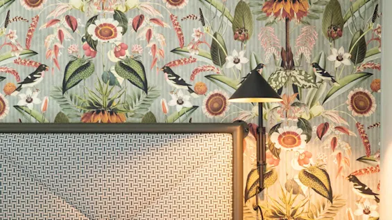 Ett mönstrat huvudgavel vilar mot en livlig tropisk tapet med stora blommor och fåglar. En svart vägglampa ger mjukt ljus i det mysiga sovrumshörnet.